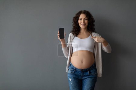 Foto de Gran aplicación. Mujer embarazada sonriente mostrando un teléfono inteligente en blanco y señalándolo, Feliz joven embarazada publicidad moderna aplicación móvil para el seguimiento del embarazo, Fondo gris, Mockup - Imagen libre de derechos