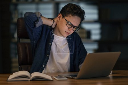 Foto de Feeling exhausted, overworked. Frustrated young handsome man in eyeglasses looks tired while sitting at his working place at dark office, using laptop, rubbing his neck, suffering from office syndrome - Imagen libre de derechos