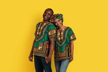 Foto de Cheery happy loving black husband and wife in beautiful traditional african costumes posing together on yellow studio background, holding hands, smiling at camera. Love, matrimony, relationships - Imagen libre de derechos