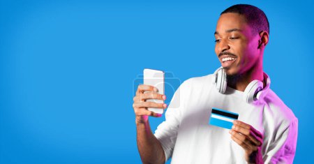Téléchargez les photos : E-commerce. Happy handsome millennial black guy in white longsleeve using modern cell phone, wireless headphones and bank credit card over studio background in neon light, panorama with copy space - en image libre de droit