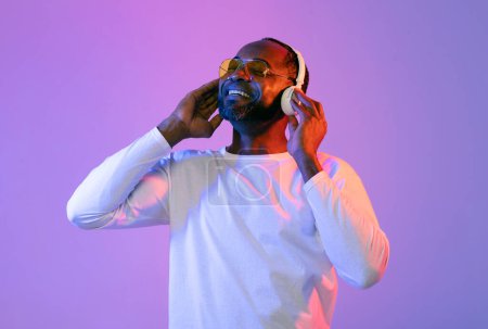 Téléchargez les photos : Joyeux homme mûr afro-américain en blanc écoutant de la musique avec les yeux fermés, utilisant des écouteurs stéréo sans fil modernes, portant des lunettes de soleil, fond de studio néon, espace de copie - en image libre de droit