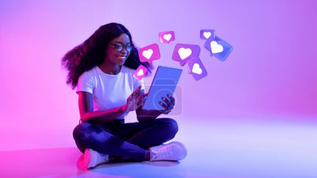 Téléchargez les photos : Glad shocked young african american lady in glasses has romantic chat with hearts on tablet on neon studio background, panorama. Dating app remotely, communication, social distance, love, new normal - en image libre de droit