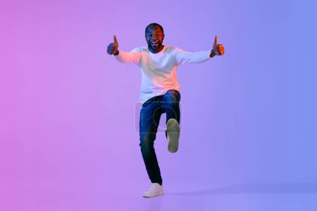 Photo for Cool happy handsome mature african american man in casual outfit showing thumb ups and shoe sole on colorful neon studio background, copy space for advertisement, full length - Royalty Free Image