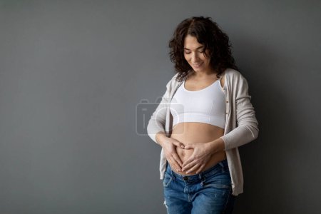 Téléchargez les photos : Beautiful Young Pregnant Woman Making Heart Gesture With Hands Near Her Belly, Smiling Expectant Female Placing Palms On Tummy And Doing Love Sign While Standing Over Grey Background, Copy Space - en image libre de droit