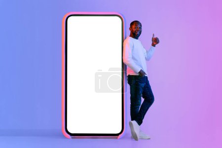 Téléchargez les photos : Positive middle aged black man in stylish outfit standing by big cell phone with white blank screen, showing thumb up, recommending nice online offer, mockup, neon studio background, full length - en image libre de droit