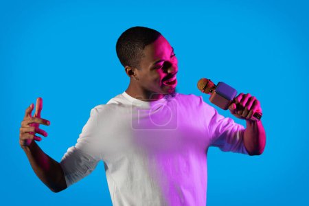 Photo for Cool attractive young black guy in casual rapper singer performing on blue studio background in neon light, talented african american man singing and gesturing - Royalty Free Image
