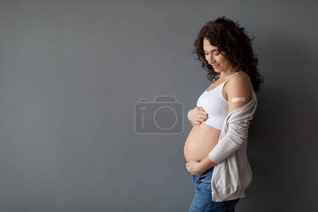 Foto de Vacunación durante el embarazo. Mujer embarazada feliz con vendaje adhesivo en el hombro abrazando su vientre mientras estaba parada sobre el fondo del estudio gris, la mujer embarazada se vacunó, espacio de copia - Imagen libre de derechos