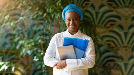 Téléchargez les photos : Pretty happy young black woman wearing traditional turban posing at cafe, smiling at camera, holding digital pad, notebook, panorama with copy space. Millennials lifestyle concept, career, education - en image libre de droit