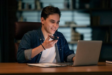 Junger gutaussehender Kerl in lässiger Arbeit spät im Büro, am Schreibtisch sitzend, mit modernem Laptop und Smartphone, Sprachnachricht aufzeichnend und lächelnd, mit Kommunikations-Handy-App, Kopierraum