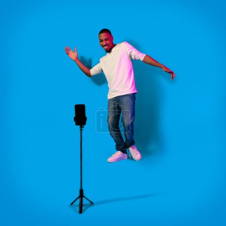 Foto de Famoso afroamericano influencer cool millennial guy grabación de contenido para las redes sociales, mirando el teléfono celular en trípode, bailando y sonriendo sobre fondo azul en luz de neón, longitud completa - Imagen libre de derechos