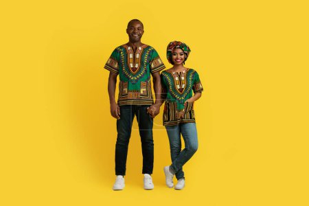 Photo for Happy cheerful beautiful traditional loving black couple in national african costumes holding hands and smiling at camera, posing together on yellow studio background, full length, copy space - Royalty Free Image