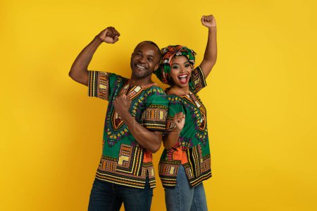 Foto de Excited happy cheerful black lovers man and woman in traditional african clothing celebrating success together on yellow studio background, standing back to back and gesturing, copy space - Imagen libre de derechos