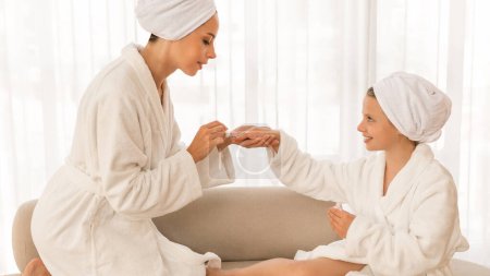 Téléchargez les photos : First Manicure Concept. Caring Mother Applying Nail Polish To Her Cute Little Daughter, Mom And Female Child Wearing White Bathrobes Having Beauty Day At Home, Enjoying Self-Care Routine - en image libre de droit