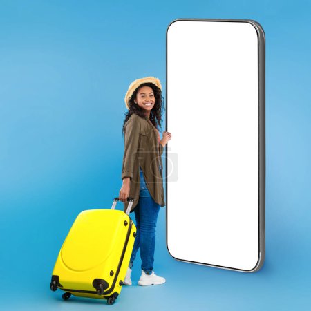 Photo for Full length portrait of happy black female traveler walking with suitcase over blue studio background, copy space. Woman tourist checking travelling online offer, standing by huge cell phone, mockup - Royalty Free Image