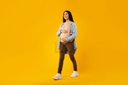 Foto de Happy pregnancy concept. Pregnant woman walking and touching belly over yellow studio background, full length shot of positive expectant lady, free space - Imagen libre de derechos