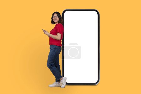 Photo for Happy pretty millennial arab woman in comfy outfit standing by big cell phone with white empty screen, using smartphone, using cool mobile app, yellow studio background, mockup, copy space - Royalty Free Image
