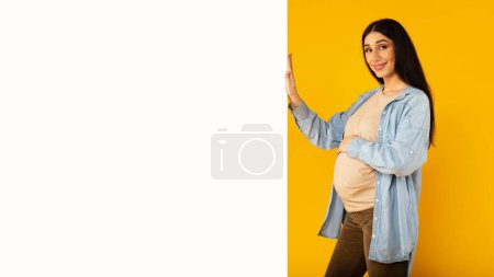 Téléchargez les photos : Joyeux jeune femme enceinte posant près du tableau blanc vierge pour le texte et toucher le ventre, la publicité quelque chose, debout sur fond de studio jaune, maquette - en image libre de droit
