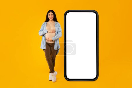 Foto de Una maqueta móvil. Mujer embarazada posando cerca de un gran teléfono inteligente con pantalla en blanco y abrazando su vientre, mujer embarazada publicidad oferta en línea o aplicación de embarazo, collage - Imagen libre de derechos