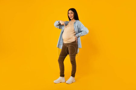 Photo for Full body length shot of pregnant woman pointing at her big belly, showing tummy and smiling, enjoying healthy pregnancy and upcoming motherhood, yellow background, free space - Royalty Free Image