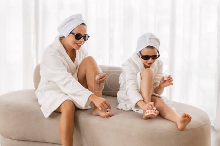 Téléchargez les photos : Mother And Little Daughter Wearing White Bathrobes And Sunglasses Doing Pedicure At Home, Happy Young Mom And Female Child Applying Nail Polish On Legs, Enjoying Domestic Beauty Day, Closeup - en image libre de droit