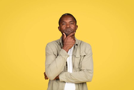 Photo for Pensive serious confident adult black male in casual thinking, presses hand to chin, isolated on yellow background, studio, free space. Choice emotions, facial expression, lifestyle, ad and offer - Royalty Free Image
