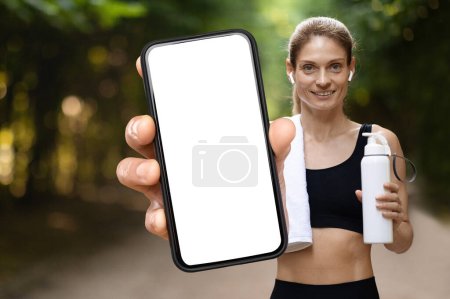 Photo for Fitness App. Sporty Middle Aged Woman Holding Big Blank Smartphone In Hand, Fit Lady Holding Water Bottle And Demonstrating Phone With White Screen, Recommending New Mobile Application, Mockup - Royalty Free Image
