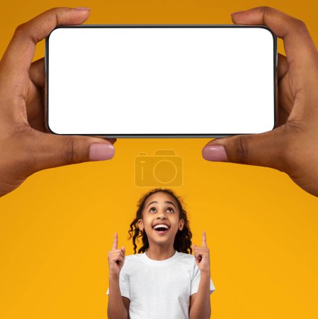 Téléchargez les photos : Nice Offer. Excited Black Girl Pointing At Huge Blank Smartphone In Giant Female Hands While Standing Isolated On Yellow Background, African American Female Child Recommending Mobile App, Mockup - en image libre de droit