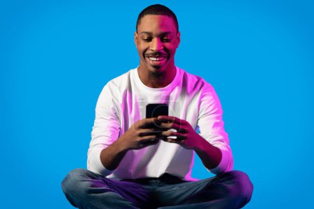 Téléchargez les photos : Joyeux jeune homme noir heureux dans occasionnel en utilisant un téléphone portable moderne et souriant sur fond bleu dans la lumière du néon, bavarder avec les dames sur sortir ensemble app, surfer sur Internet. Concept de dépendance au gadget - en image libre de droit