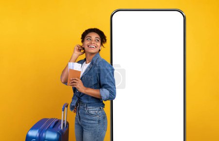 Photo for Travelling App Concept. Cheerful African American Woman Tourist With Passport, Tickets And Luggage Standing Ready For Vacation Posing On Yellow Studio Background Next To Big Phone, Mockup, Collage - Royalty Free Image
