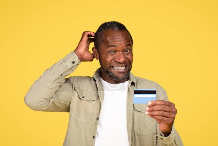 Téléchargez les photos : Confused mature black male in casual scratching head, thinking, show credit card, isolated on yellow background, studio. Sale choice, problems with shopping, advertising and offer, facial expression - en image libre de droit
