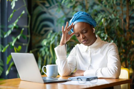 Photo for Unhappy tired frustrated young black woman entrepreneur working online from coffee shop, suffering from headache, touching head, feeling bad, using computer notebook and earpods, copy space - Royalty Free Image