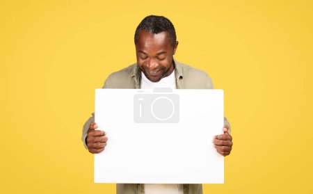 Téléchargez les photos : Heureux mâle noir d'âge moyen en tenue décontractée et regarder grande bannière avec espace vide pour le texte, isolé sur fond jaune, studio. Annonce et offre, recommandation de vente, conseils professionnels, nouvelles énormes - en image libre de droit