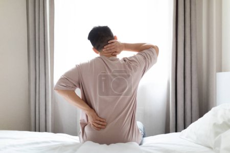 Photo for Tired man suffering from back pain after sleeping in bedroom, waking up in morning, sitting on bed and touching lower back and neck, experiencing body aches, wearing pajamas, copy space, back view - Royalty Free Image