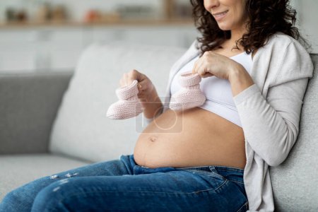Photo for Birth Awaiting. Pregnant lady sitting with little baby shoes in hands, smiling young expectant woman placing small knitted boots on her belly while resting on couch in living room at home, copy space - Royalty Free Image