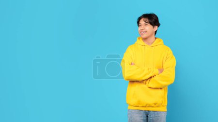 Photo for Happy Chinese Teen Boy Looking Aside At Free Space For Text Standing Crossing Hands Over Blue Studio Background, Wearing Yellow Hoodie. Male Teenager Posing Advertising Great Offer. Panorama - Royalty Free Image