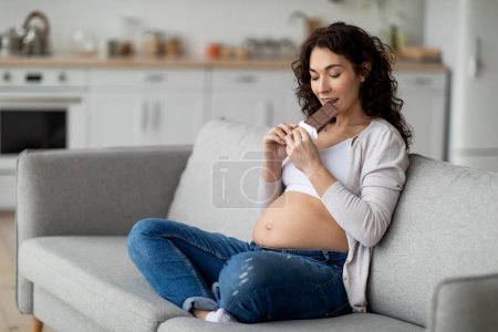 Téléchargez les photos : Pregnancy Cravings. Young pregnant woman eating bar of chocolate at home, hungry expectant lady enjoying sugary treat while relaxing on couch in living room, choosing unhealthy foods, copy space - en image libre de droit