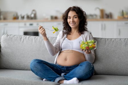 Photo for Happy Young Pregnant Lady Eating Fresh Vegetable Salad At Home, Beautiful Expectant Woman Sitting On Couch In Living Room With Bowl Of Healthy Vitamin Food And Smiling At Camera, Copy Space - Royalty Free Image
