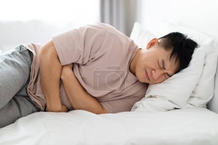 Téléchargez les photos : Unhappy sick middle aged asian man lying in bed and touching his belly, suffering from stomach pain in the morning, wearing pajamas, home interior. Gastrointestinal Diseases, Problems - en image libre de droit