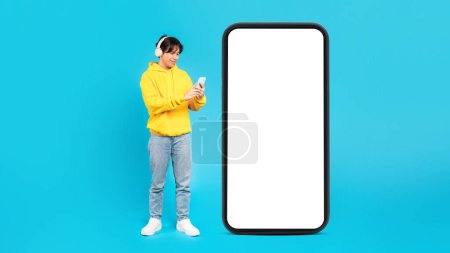 Foto de Japanese Male Teen Near Large Cellphone With Empty Screen Using Mobile Phone Wearing Headphones In Studio On Blue Background. New Application Advertisement. Panorama, Mockup - Imagen libre de derechos