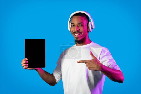 Téléchargez les photos : Musical app. Happy cheerful handsome young black man showing digital tablet with white empty screen in neon light, using modern wireless headphones, pointing at pad with mockup, blue background - en image libre de droit