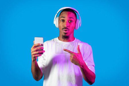 Téléchargez les photos : Choqué surpris jeune homme noir pointant vers smartphone dans sa main, en utilisant des écouteurs sans fil, écouter de la musique en ligne sur bleu dans la lumière au néon, vérifier agréable application mobile divertissante - en image libre de droit