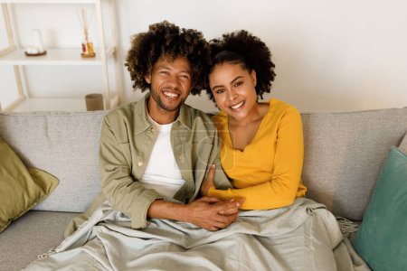 Téléchargez les photos : Joyeux couple noir embrassant sourire à la caméra assis sur le canapé posant à la maison. Conjoints Profitant Homey Week-end ensemble. Mariage heureux et concept de relation romantique - en image libre de droit