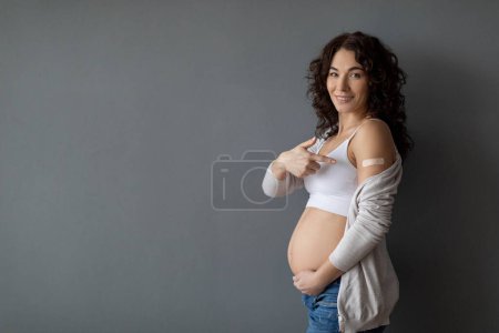 Photo for Immunization Concept. Smiling Pregnant Woman Showing Arm With Medical Bandage After Vaccine Injection, Happy Expectant Lady Pointing At Shoulder With Plaster, Standing On Grey Background, Copy Space - Royalty Free Image