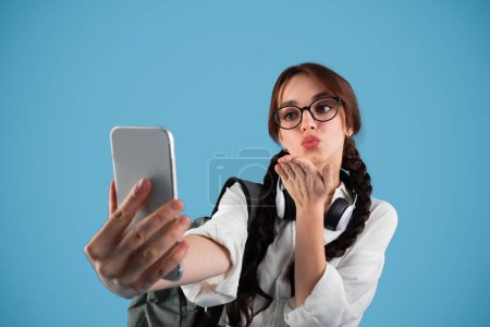 Téléchargez les photos : Smiling teen european girl with pigtails and backpack in glasses and headphones makes photo on phone making blowing kiss isolated on blue studio background. Tech, app for blog and social networks - en image libre de droit