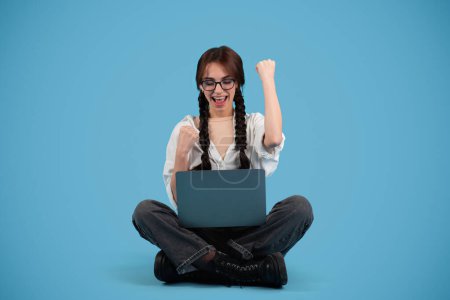 Téléchargez les photos : Happy teen european girl student with pigtails in glasses with laptop, rejoices to victory, make success gesture, isolated on blue background. Great news, study and knowledge at school, ad, offer - en image libre de droit
