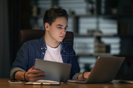 Photo for Focused young handsome man in casual outfit employee sitting at workdesk, using laptop and digital pad, working at office in evening, copy space. Modern technologies in business - Royalty Free Image