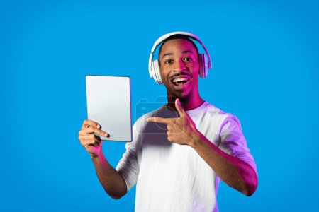 Téléchargez les photos : Happy cheerful young handsome african american guy pointing at modern digital pad, using wireless headphones and smiling on blue in neon light, listening to music, gaming online, watching video - en image libre de droit