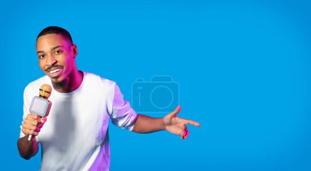 Téléchargez les photos : Cool handsome young african american guy in casual hip hop singer performing on blue studio background in neon light, black man singing and gesturing, panorama with copy space - en image libre de droit