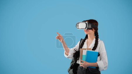 Téléchargez les photos : Smiling teen european girl with pigtails and backpack in vr glasses controls air with hand, point finger at copy space, isolated on blue studio background. Gadget for study and knowledge, education - en image libre de droit