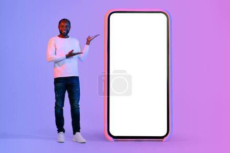 Téléchargez les photos : Cheerful middle aged black man in stylish outfit pointing at big cell phone with white blank screen, showing nice online offer, mockup, copy space, neon studio background, full length - en image libre de droit